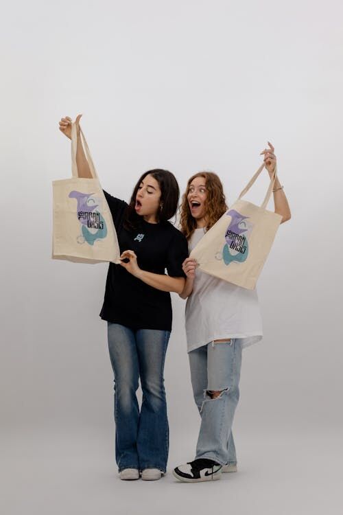 Free Two women joyfully display eco-friendly tote bags with creative design on a white background. Stock Photo